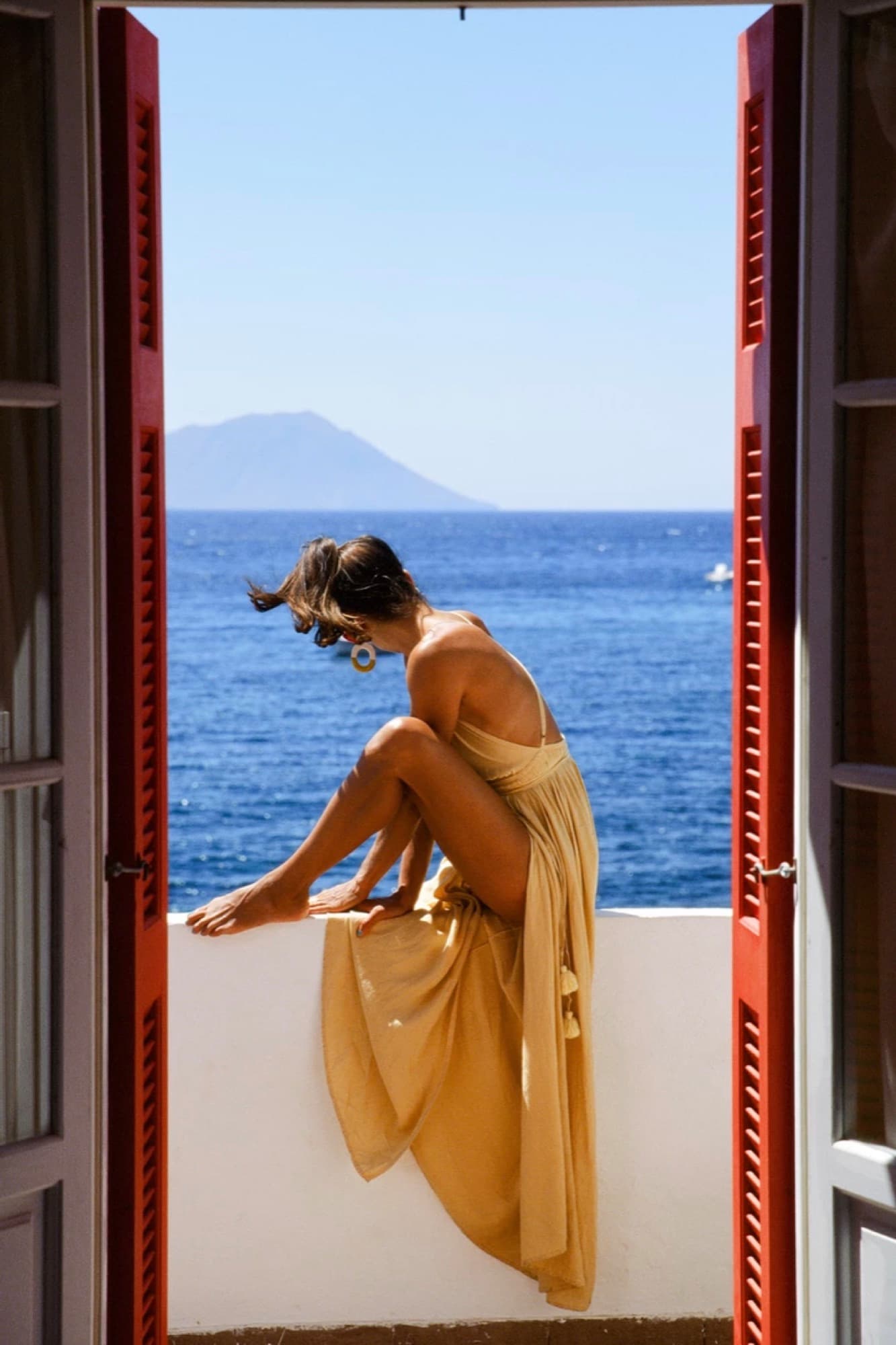 A woman in a gold dress perched in an open balcony door above the sea