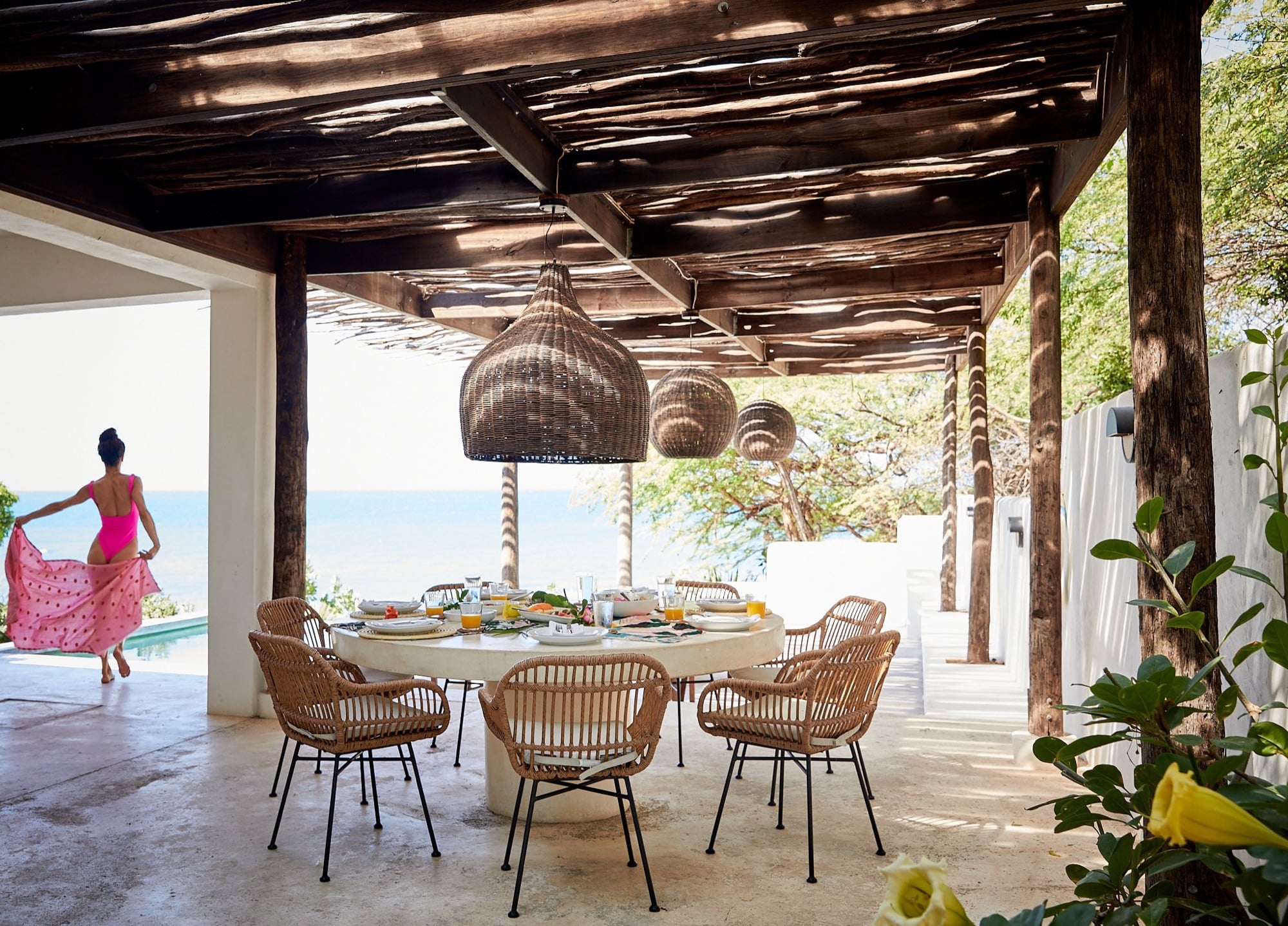 A woman in a pink sarong runs across a shaded resort veranda set for lunch